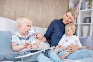 Mutter hat ein kleines Kind auf dem Schoß, das ältere Kind sitzt daneben und inhaliert mit Vernebler