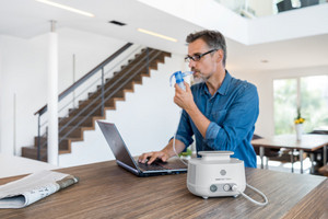 Man sitting at table in front of laptop while inhaling with inhalation device
