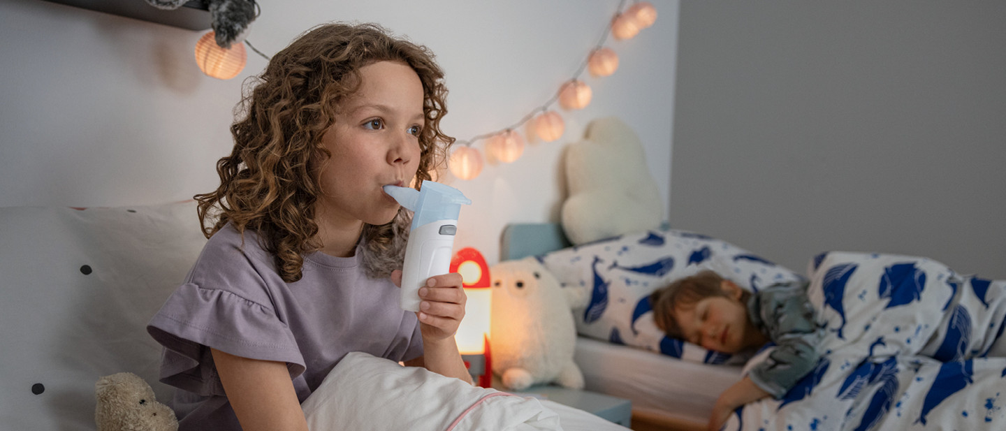 Ein kleines Mädchen sitzt im Bett und inhaliert mit einem PARI BOY Go. Neben ihr schläft ruhig ihr Bruder.