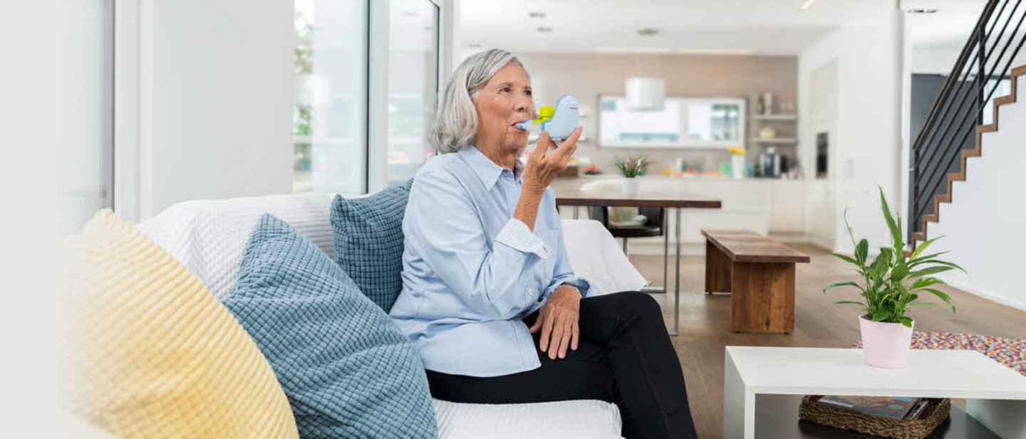Ältere Dame sitzt auf weißem Sofa und benutzt ein PARI Inhaliergerät in heller, moderner Wohnung mit Wohnzimmer, Küche und Treppe im Hintergrund.