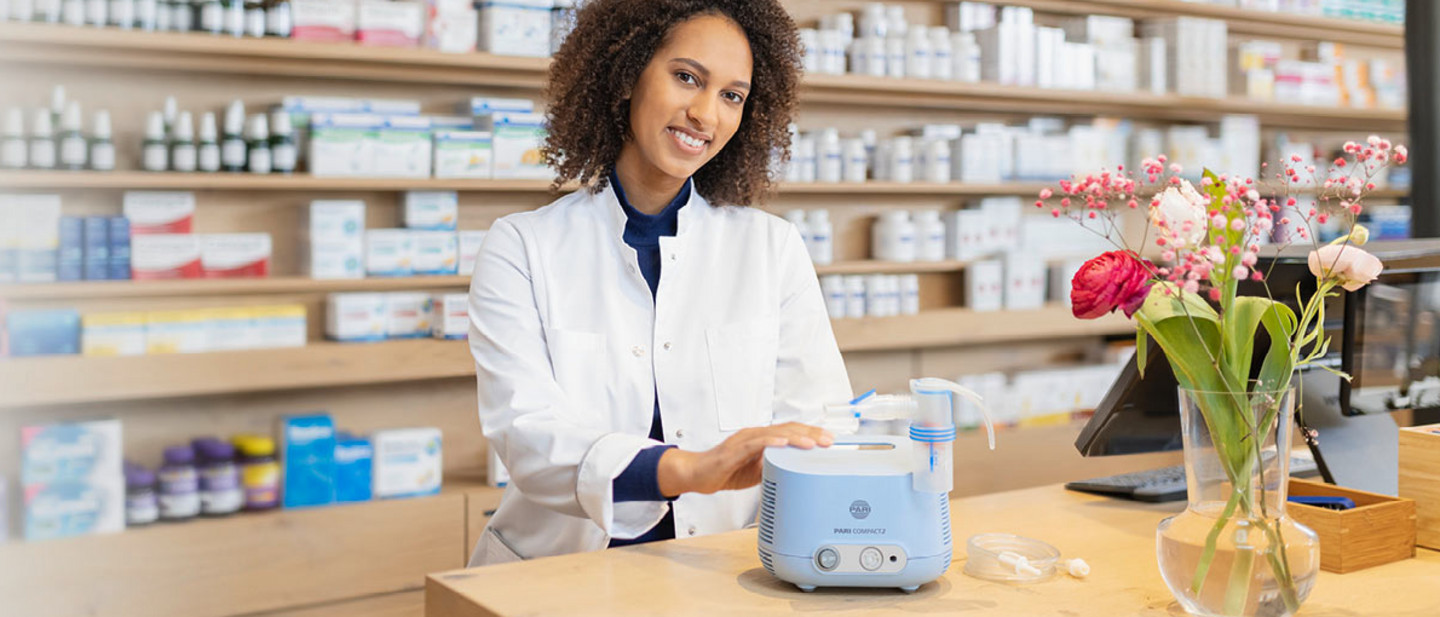 In einer Apotheke steht eine Apothekerin hinter der Theke und erklärt die Funktionsweise des PARI COMPACT2 Inhaliergerätes, das vor ihr auf der Theke steht. Lächelnd legt sie dabei die rechte Hand auf das Gerät. Rechts neben ihr steht eine Vase mit Blumen. Die Apotheke ist modern, hell und freundlich.
