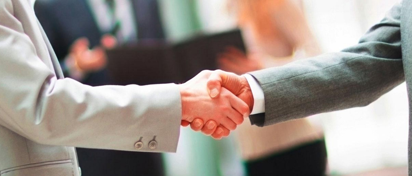 Zwei Personen in Anzügen geben sich vor unscharfem Hintergrund die Hand, im Raum stehen weitere Personen.