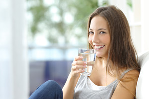 Lächelnde, junge Frau sitzt mit Glas Wasser in der Hand auf dem Sofa