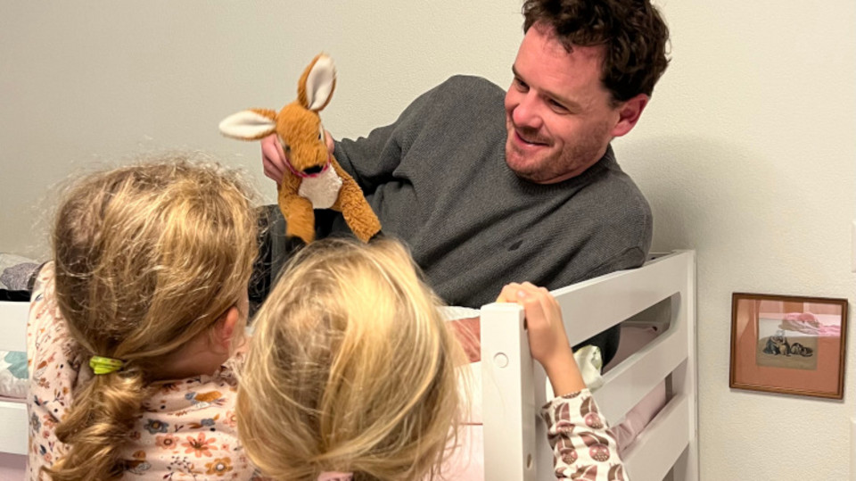 Vater liegt auf Hochbett mit Kuscheltier in der Hand, seine Töchter stehen davor und schauen ihm zu