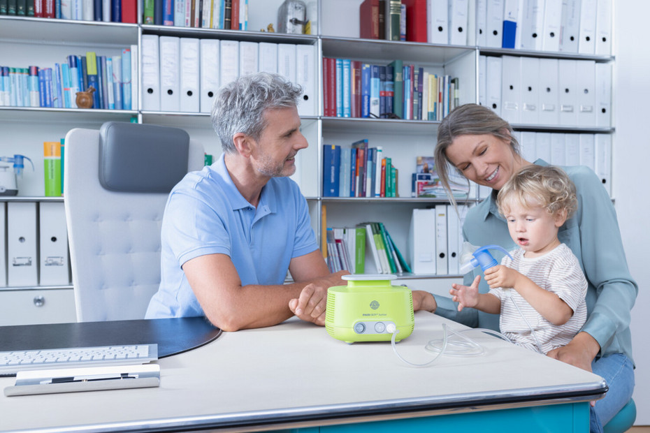 Mutter sitzt mit ihrem Kind beim Kinderarzt, der beiden ein Inhalationsgerät zeigt