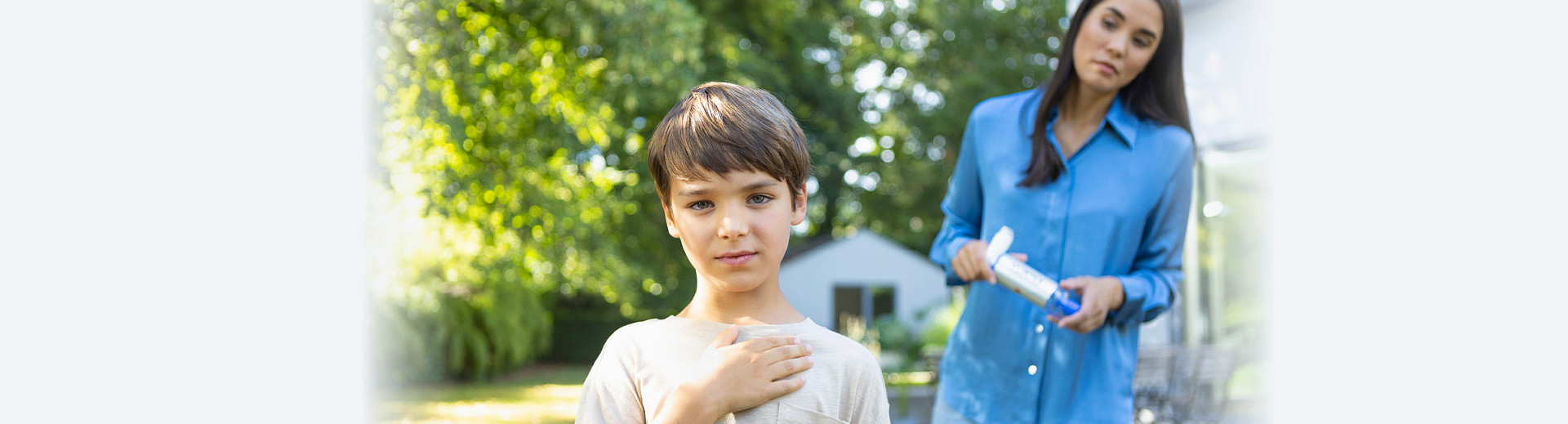 Ein Junge steht mit einer Hand auf der Brust im Garten, während eine Frau in einer blauen Bluse mit einer VORTEX Inhalierhilfe in der Hand besorgt hinter ihm steht.
