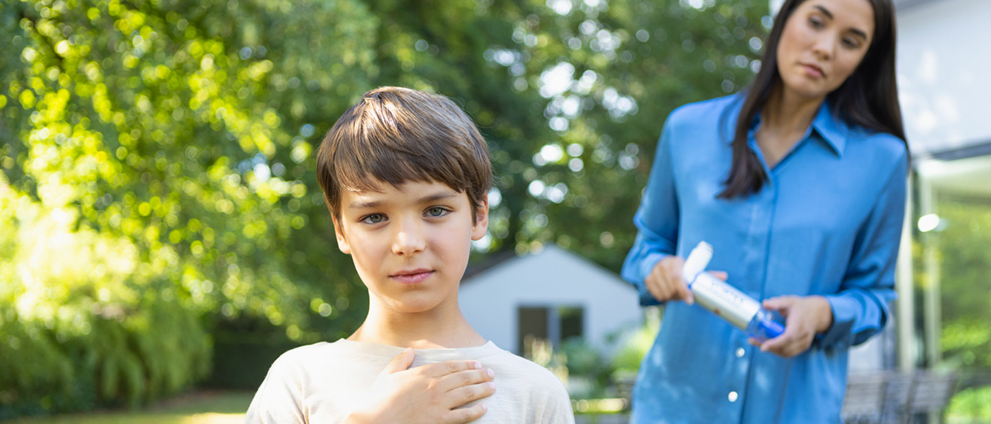Junge steht im Garten und hält eine Hand an die Brust, während seine Mutter im Hintergrund ein Asthmaspray mit Vorschaltkammer hält.