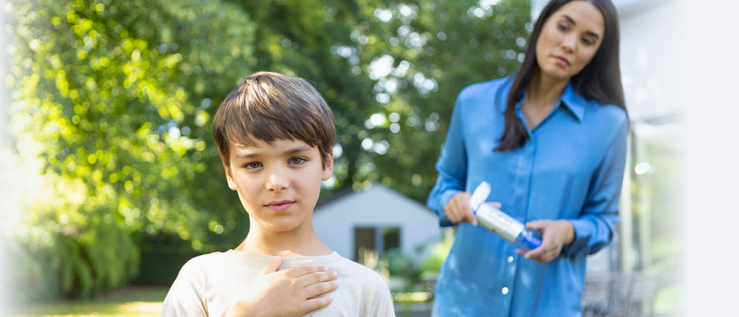 Ein Junge steht mit einer Hand auf der Brust im Garten, während eine Frau in einer blauen Bluse mit einer VORTEX Inhalierhilfe in der Hand besorgt hinter ihm steht.