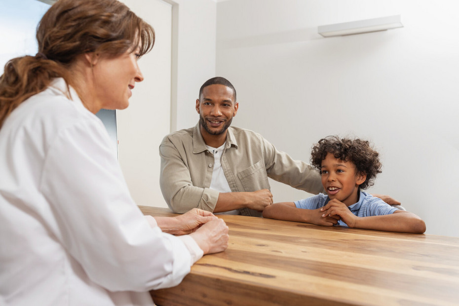 Doctor at the table in the foreground, father and son sitting in front of her