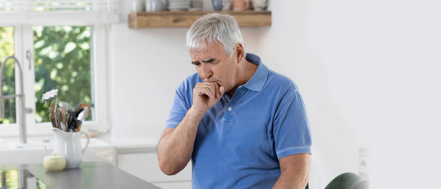An older man with gray hair and a blue polo shirt stands in a bright kitchen and coughs into his hand.