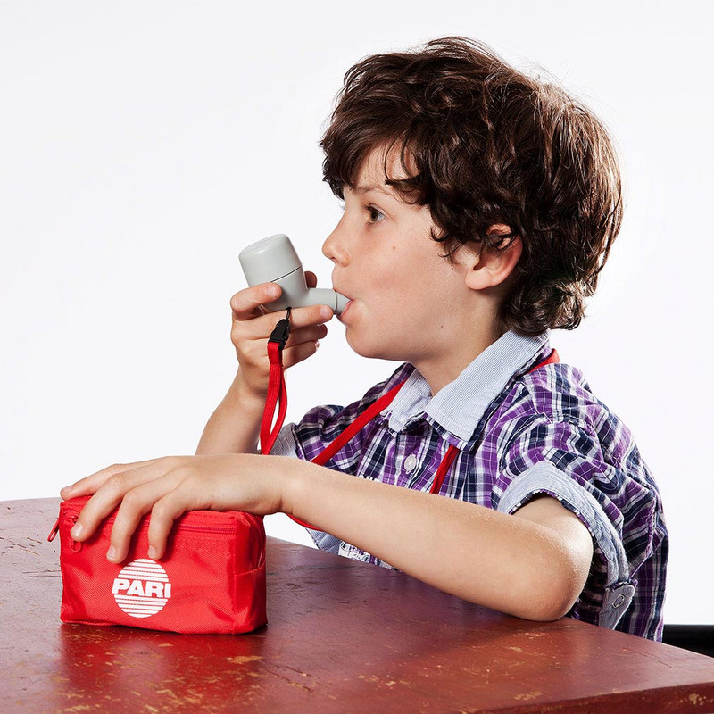 Ein Junge sitzt an einem Tisch, in aufrechter Haltung. In der rechten Hand hält der das PARI O-PEP, dessen Mundstück er mit seinen Lippen umschließt.