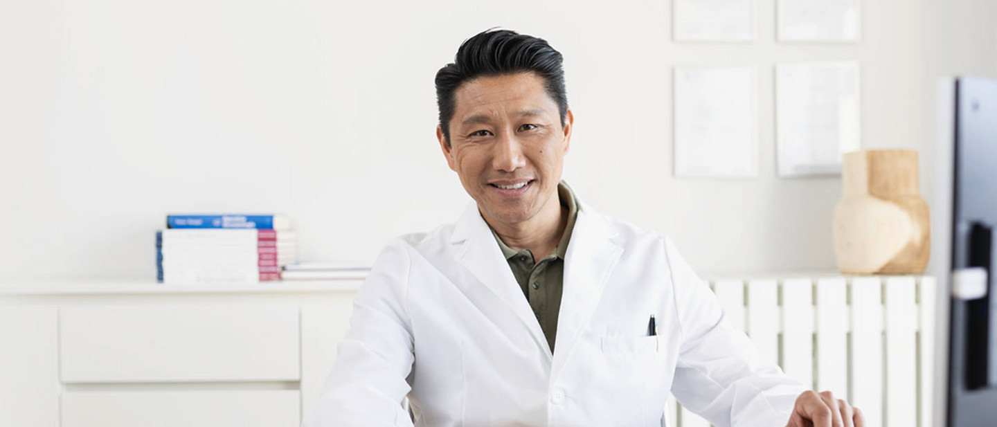 Freundlich lächelnder Arzt in weißem Kittel sitzt an einem Schreibtisch und arbeitet an einem Computer in einer modernen Praxis.