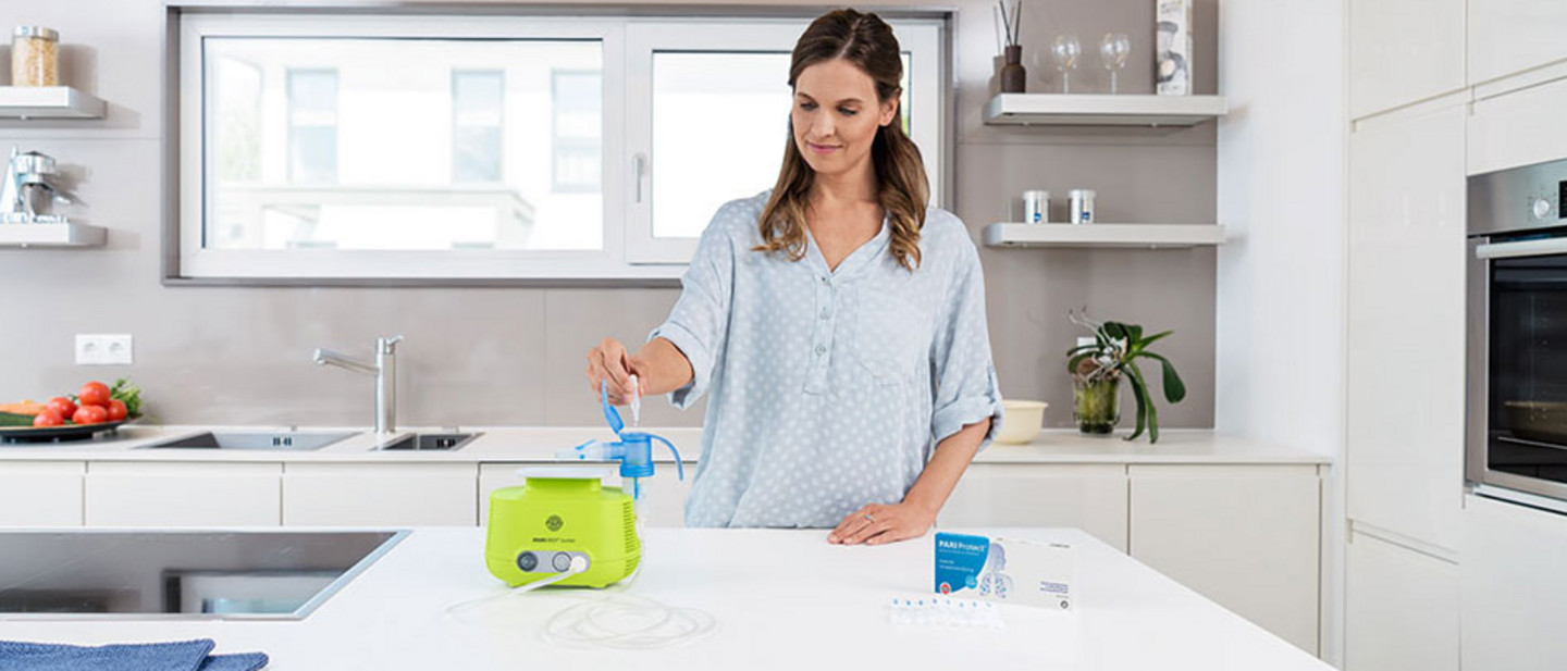Dans une cuisine moderne, une femme remplit le nébuliseur d'un PARI BOY Junior avec une solution pour inhalation PARI Protect.Dans une cuisine moderne, une femme remplit le nébuliseur d'un PARI BOY Junior avec une solution pour inhalation PARI Protect.