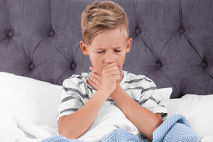 Boy sitting in bed coughing, holding one hand to his mouth, the other hand to his throat