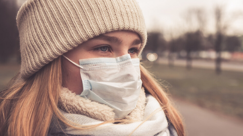 Young woman wearing a scarf, hat and face mask standing at a bus stop