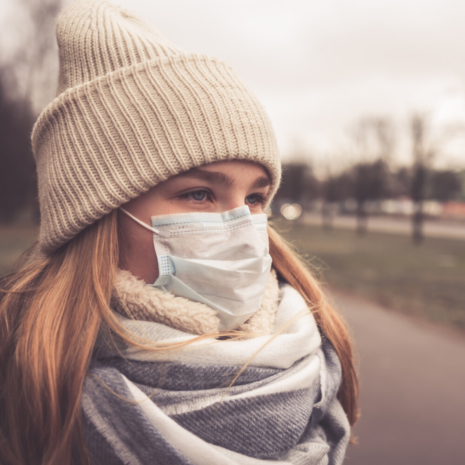 Young woman wearing a hat and scarf stands at a bus stop wearing a face mask