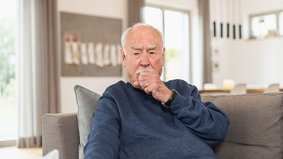 Elderly man sits on the couch and breathes through his fist