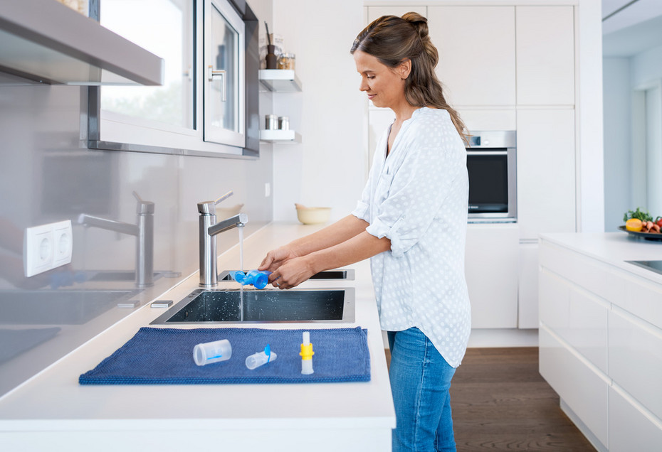 A woman stands in front of the kitchen sink, cleaning parts of the disassembled nebulizer under running water. The already cleaned nebulizer parts lie on a cloth next to the sink to dry.