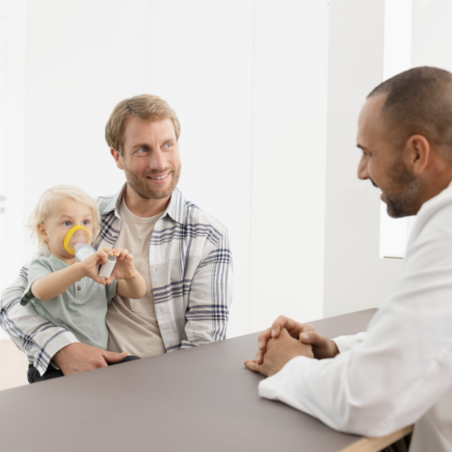 Junger Vater sitzt mit seinem kleinen Sohn beim Arzt, der Sohn inhaliert Asthmaspray mit Inhalierhilfe VORTEX