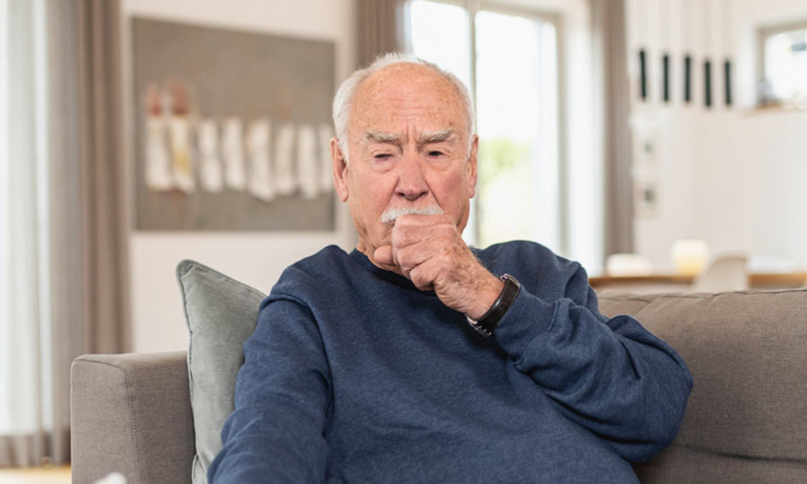 Hustender Mann mit Bronchitis Älterer Mann sitzt auf einem Sofa und hustet in seine Hand, im Hintergrund ein heller Wohnraum.