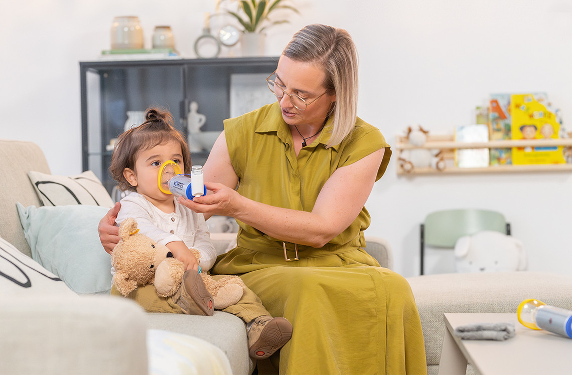 Richtige Anwendung von Asthmaspray Mutter hält ihrem Kleinkind auf dem Sofa ein Asthmaspray mit VORTEX Inhalierhilfe und gelber Babymaske zur Inhalation auf Nase und Mund