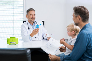 Arzt erklärt Vater und Sohn Inhaliergerät, das vor ihm auf dem Tisch steht, in der Hand hält er den Vernebler