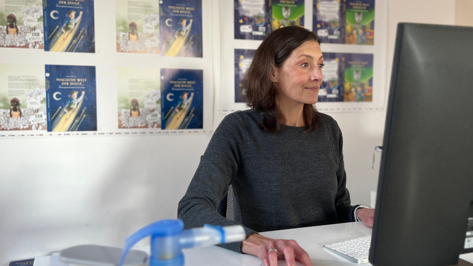 Junge Frau sitzt vor Computer am Schreibtisch, im Vordergrund ist unscharf ein Inhalationsgerät zu erkennen