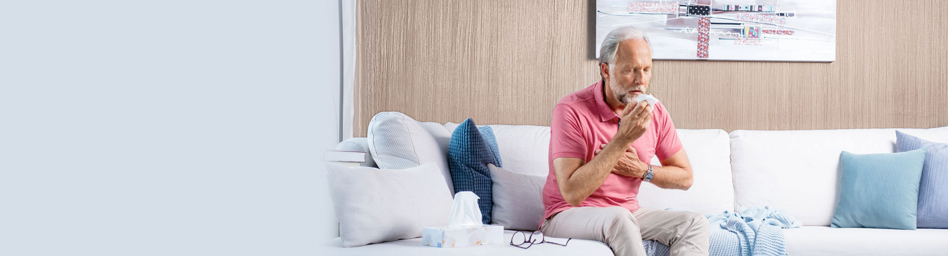 An elderly man sits on the sofa, coughing heavily and holding his hand in front of his chest