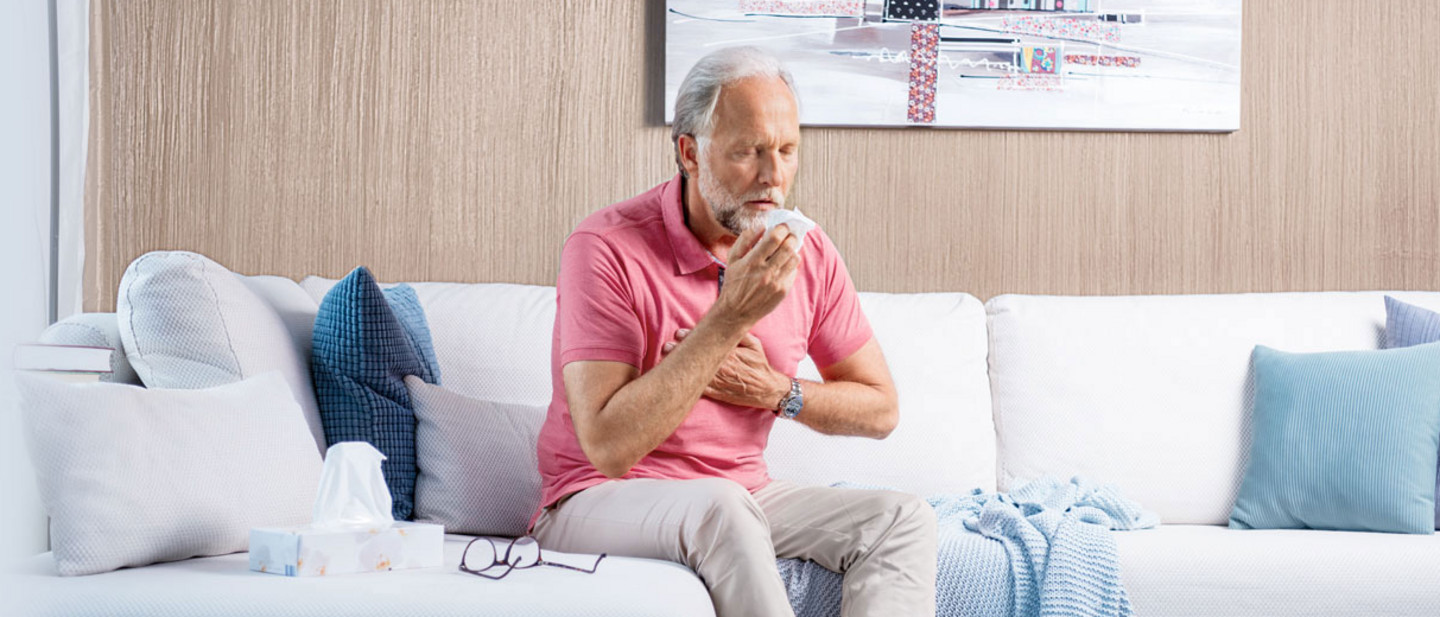 An elderly man sits on the sofa, coughing heavily and holding his hand in front of his chest