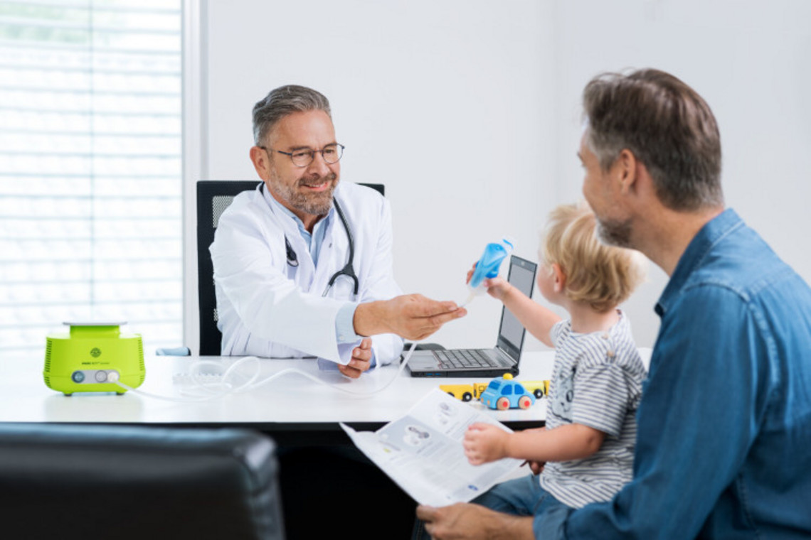 FAQ zu Inhalation bei Asthma: Beim Arzt Arzt sitzt am Schreibtisch und reicht einem Kleinkind einen blauen Vernebler, der mit einem Inhalationsgerät verbunden ist, während der Vater daneben sitzt.