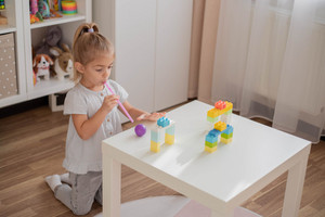 Little girl kneels in front of play table and blows balls over it with a straw