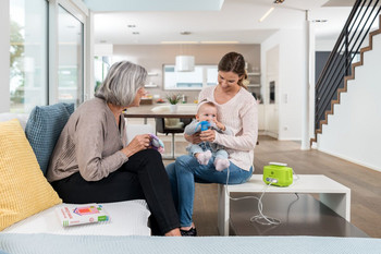 Oma sitzt auf Couch und beobachtet Mutter mit Baby auf dem Schoß, das mit dem PARI BOY Junior Inhalationsgerät inhaliert