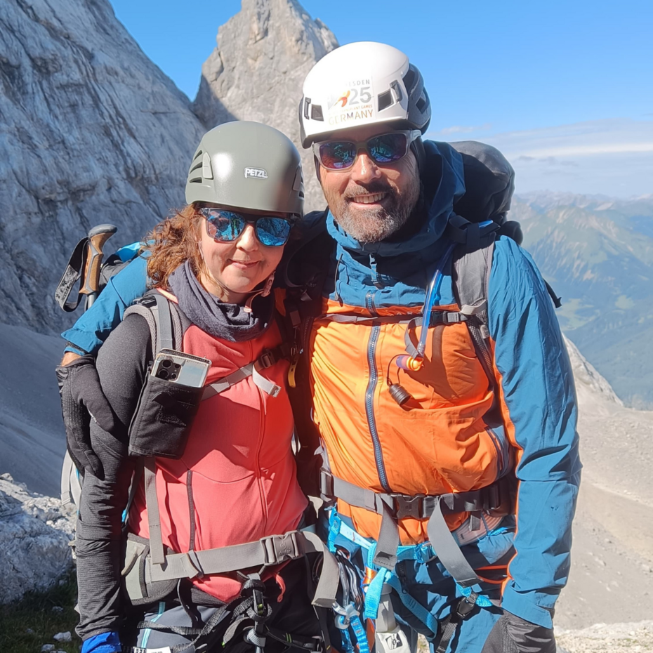 Eine Frau und ein Mann in Bergausrüstung lächeln in die Kamera, hinter ihnen ein Berg