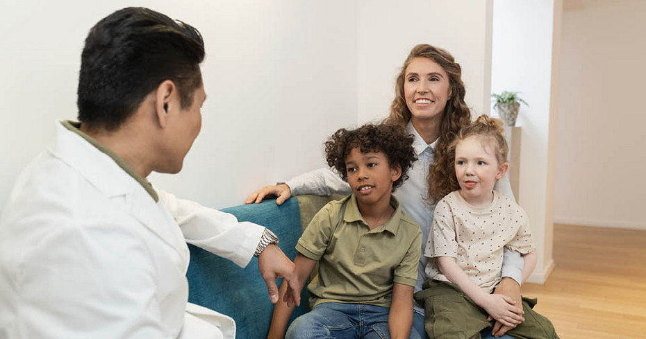 A healthcare professional sits on a chair talking to an adult with two children in a bright room.