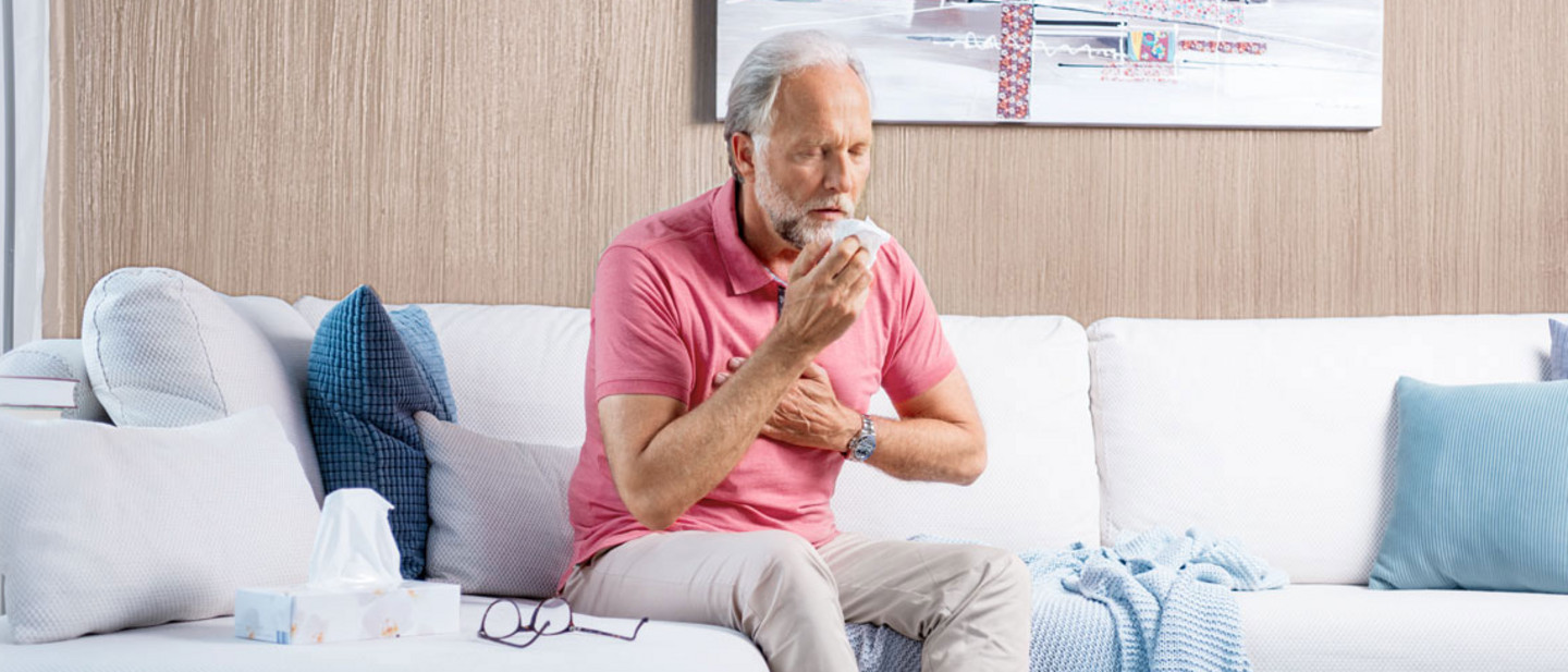 An elderly man sits on the sofa, coughing heavily and holding his hand in front of his chest