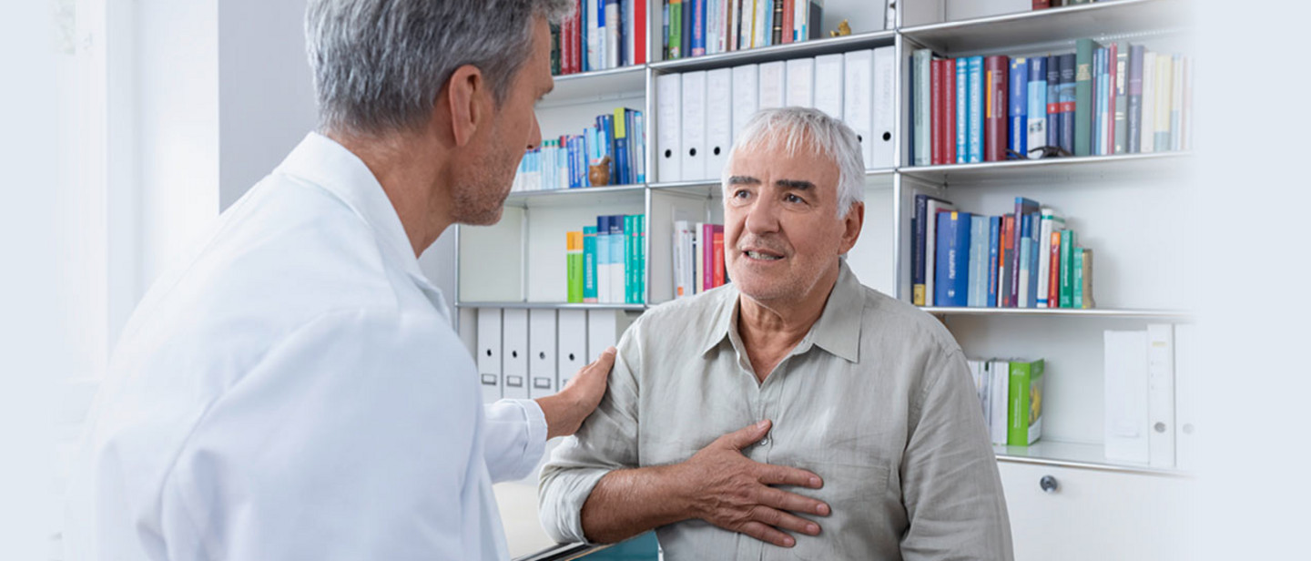 Wir sind da, wenn Luft holen nicht mehr selbstverständlich ist Ein älterer Mann sitzt in einer Arztpraxis, hält seine Hand auf die Brust und spricht mit einem Arzt im weißen Kittel, der ihm beruhigend eine Hand auf die Schulter legt.