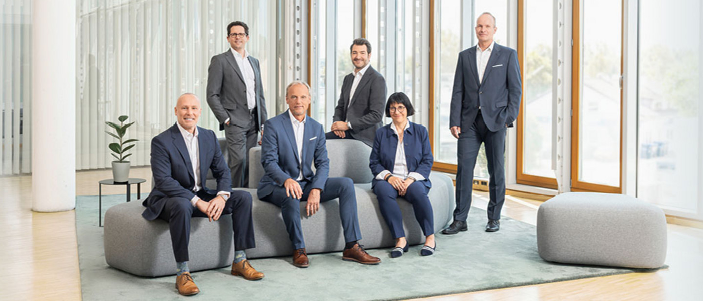 Das Management der PARI Unternehmensgruppe Fünf Männer und eine Frau in Businesskleidung stehen und sitzen in einem modernen, lichtdurchfluteten Raum auf grauen Sitzmöbeln.