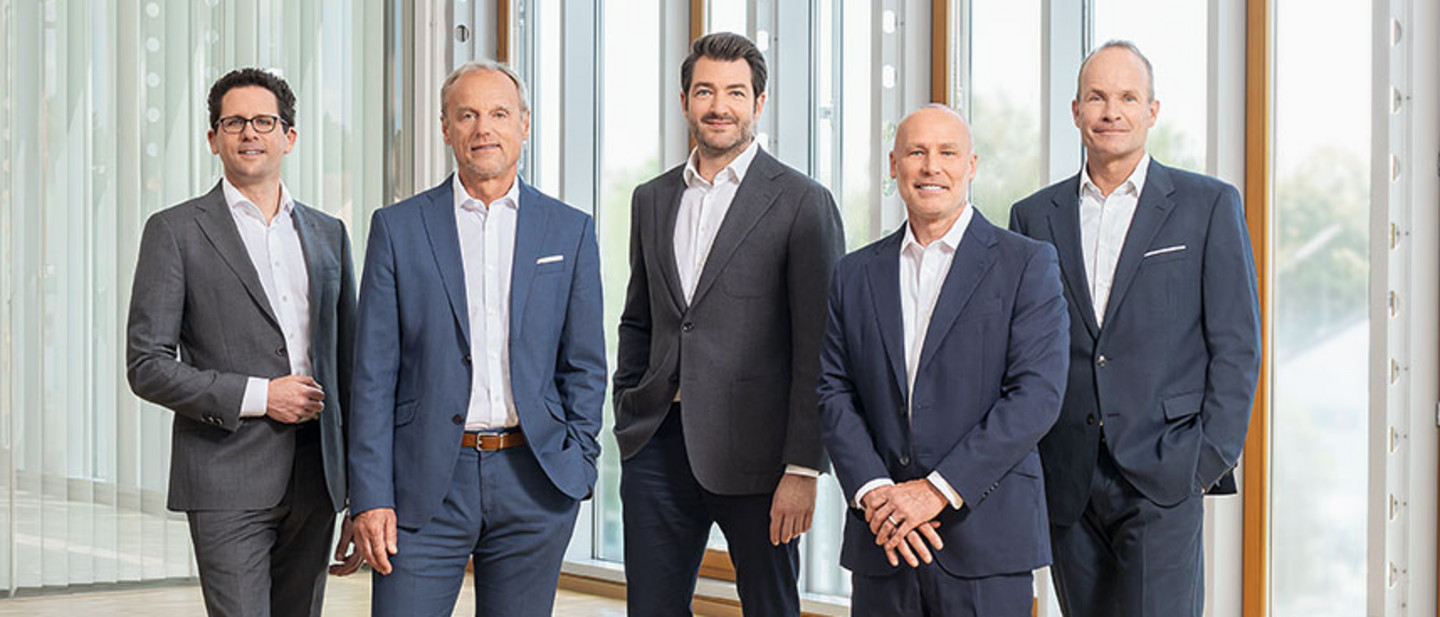 Fünf Männer und eine Frau in Businesskleidung stehen und sitzen in einem modernen, lichtdurchfluteten Raum auf grauen Sitzmöbeln.