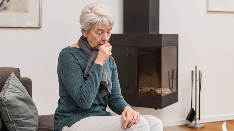 Ältere Frau sitzt im Wohnzimmer auf der Couch und hustet in die geschlossene Faust