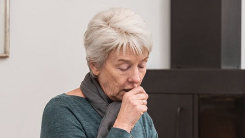 Ältere Frau sitzt im Wohnzimmer auf der Couch und hustet in die geschlossene Faust