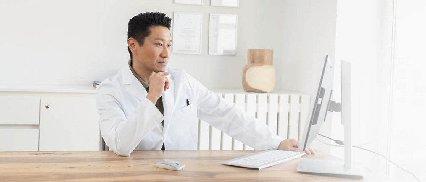A doctor in a white coat is sitting at a desk, working intently on a computer in a bright office.