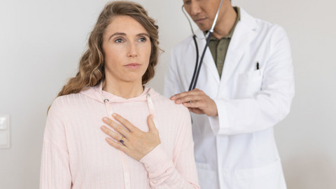 Chronische Pseudomonas-Besiedelung der Lunge bei Mukoviszidose: Ein Erfahrungsbericht Junge Frau sitzt auf einer Liege mit Hand auf der Lunge, während Arzt hinter ihr steht und sie mit Stethoskop abhört