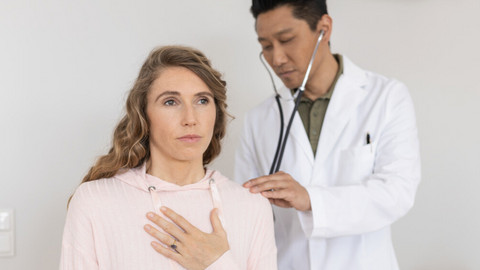 Junge Frau sitzt auf einer Liege mit Hand auf der Lunge, während Arzt hinter ihr steht und sie mit Stethoskop abhört