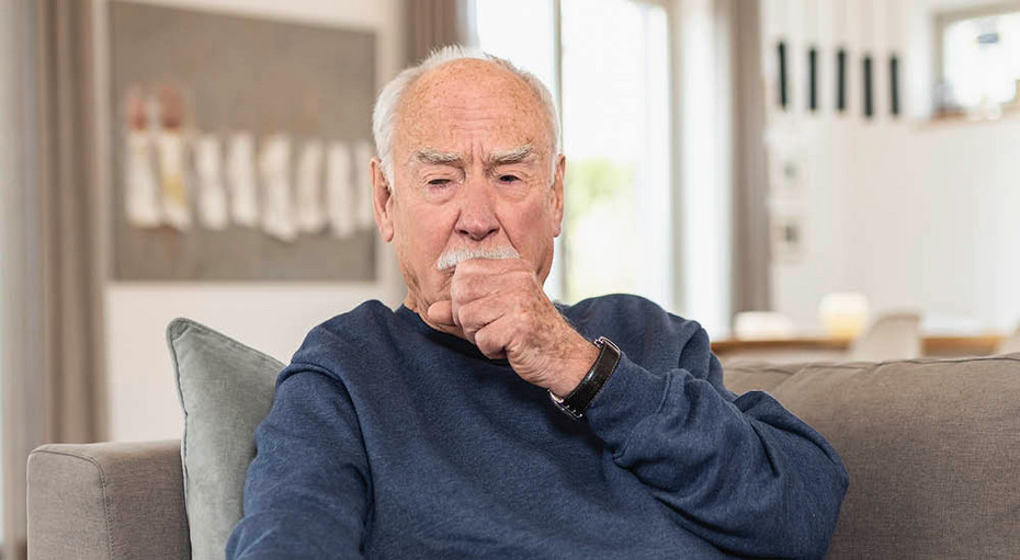 Dry cough management An elderly man sits on a sofa in a living room. He wears a dark blue sweater and has a thoughtful expression as he coughs into his hand.