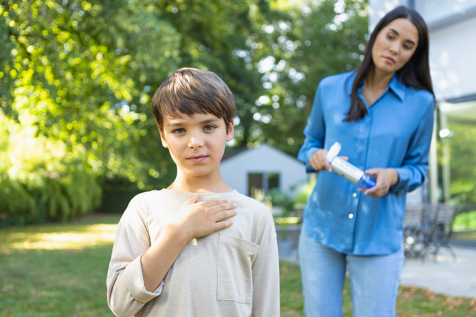 Young boy holding his hand on his chest, mother in the background bringing asthma spray with VORTEX spacer