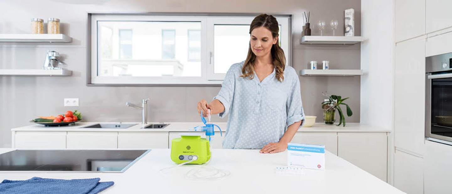 A woman fills the nebuliser of a PARI BOY Junior with PARI Protect inhalation solution in a modern kitchen.