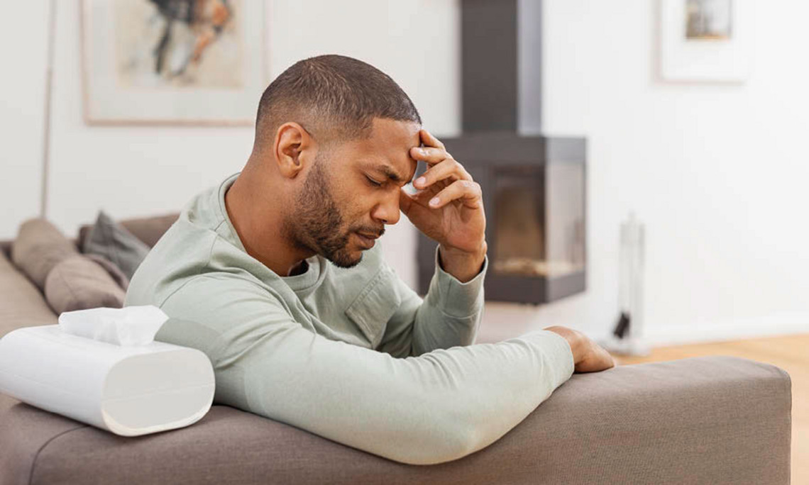 Symptome einer Sinubronchitis Mann sitzt auf einem Sofa in hellem Wohnzimmer, stützt den Kopf mit der Hand und wirkt erschöpft, neben ihm liegt eine Taschentuchbox.