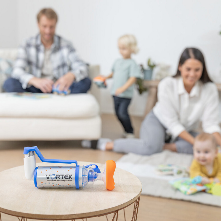 Spacer inhalation with babies: VORTEX holding chamber In the background, a young family is playing in the living room; in the foreground, the VORTEX holding chamber is lying on a small table