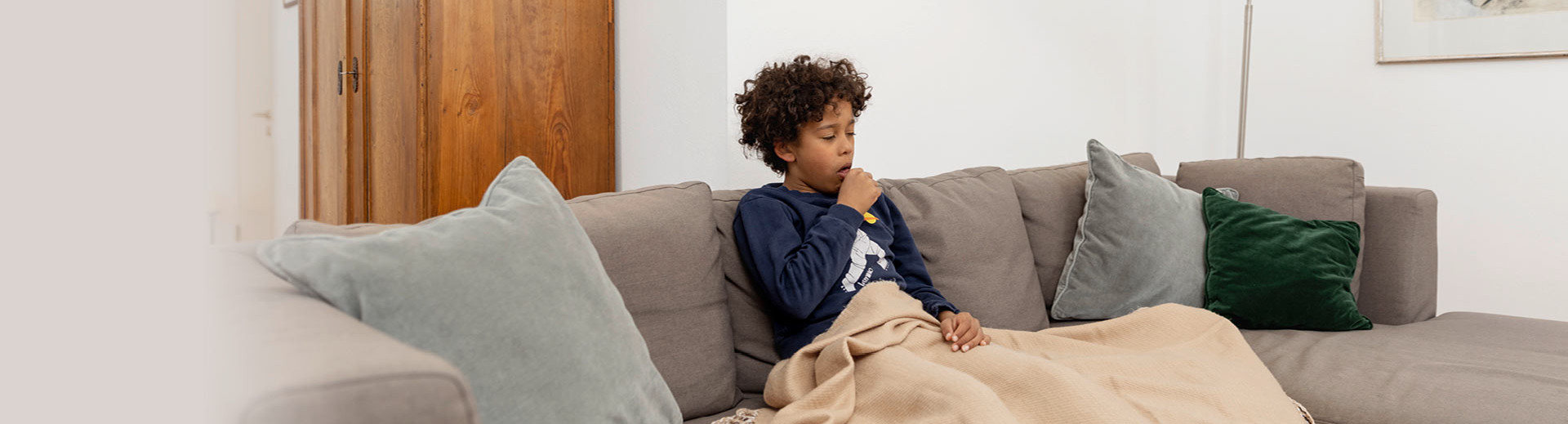 Ein kranker Junge sitzt zu Hause auf dem Sofa unter einer Decke und muss husten
