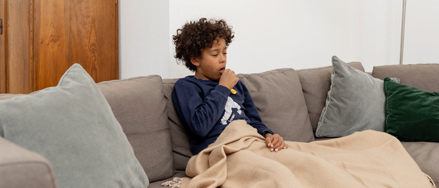 Ein Junge mit lockigen Haaren sitzt krank auf einem grauen Sofa, eingewickelt in eine beige Decke. Er hustet in seine Hand.